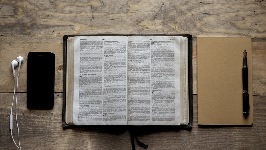 Aufgeschlagene Bibel auf einer Holzoberfläche Geöffnete Bibel; rechts daneben ein Notizbuch, auf dem ein Füller liegt. Links von der Bibel liegt ein ein Smartphone mit einem Kopfhörer.