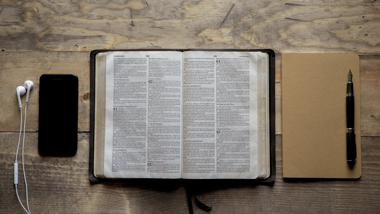 Geöffnete Bibel; rechts daneben ein Notizbuch, auf dem ein Füller liegt. Links von der Bibel liegt ein ein Smartphone mit einem Kopfhörer.
