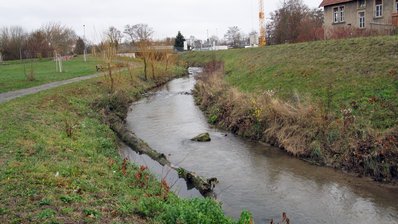 Der Leimbach in einem Abschnitt, in dem dieser bereits in einen naturnahen Zustand gebracht wurde: mit leicht mäandrierendem Gewässerlauf, Steinen im Gewässer, die für vielfältige Strömungsverhältnisse sorgen und seitlichem Bewuchs. Gewässerbegleitend verläuft ein Uferweg mit einer Bank. Im Hintergrund sieht man eine Brücke, einen Kran und ein Gebäude. 