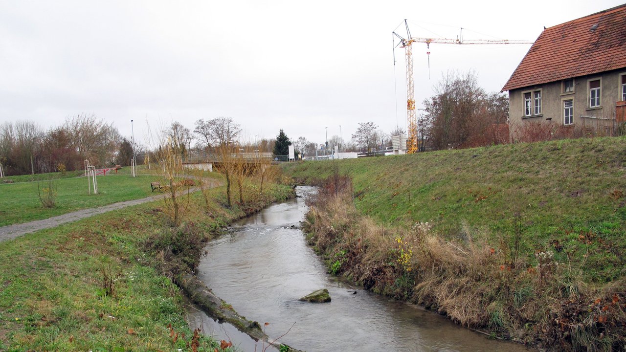 Der Leimbach in einem Abschnitt, in dem dieser bereits in einen naturnahen Zustand gebracht wurde: mit leicht mäandrierendem Gewässerlauf, Steinen im Gewässer, die für vielfältige Strömungsverhältnisse sorgen und seitlichem Bewuchs. Gewässerbegleitend verläuft ein Uferweg mit einer Bank. Im Hintergrund sieht man eine Brücke, einen Kran und ein Gebäude. 