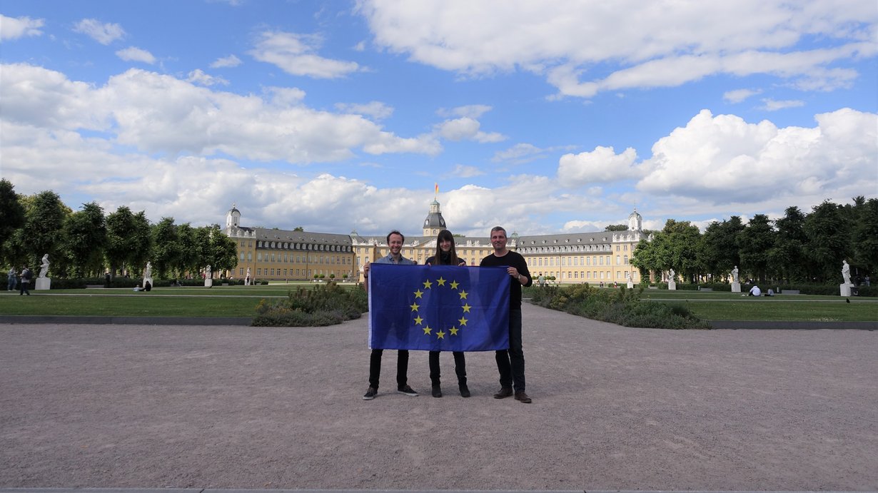 Im Vordergrund drei Mitglieder mit der EU Flagge bedruckt. Im Hintergrund ist unter blauem Himmel das Karlsruher Schloss zu sehen. 
