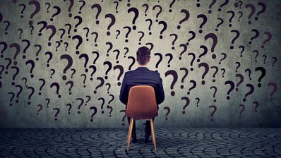 Mann, der auf einem Stuhl vor einer grauen Wand sitzt, auf der viele Fragezeichen angebracht sind