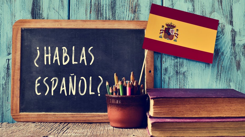 Kreidetafel mit Beschriftung "hablas espanol?" Kreidetafel mit Beschriftung "hablas espanol?" vor türkisfarbener Wand. Davor ein Gefäß mit Stiften und spanischer Fahne. Rechts neben dem Gefäß liegen zwei übereinander gestapelte Bücher.