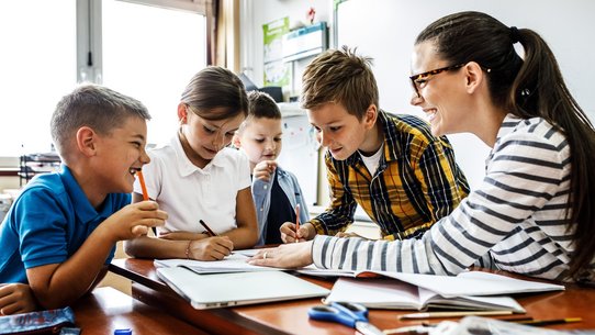 Eine Lehrerin lernt mit 4 Kindern an einem Tisch, Bücher, Hefte und Stifte liegen darauf