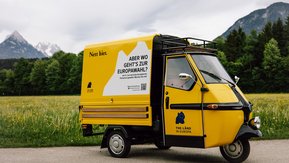 mittig eine gelbe Kaffee-Ape mit der Aufschrift „Nett hier. Aber wo geht’s zur Europawahl“ sowie dem Logo „The Länd in Europa“ auf der Fahrertür.