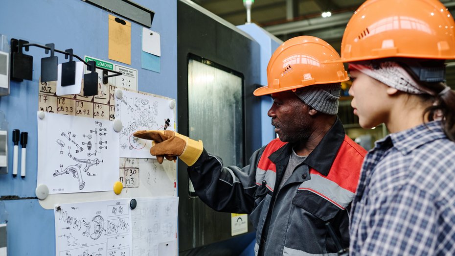 Ausländische Fachkräfte Zwei ausländische Fachkräfte untersuchen detaillierte, an eine Pinnwand gepinnte Baupläne und diskutieren wichtige Aspekte der technischen Zeichnungen in einem industriellen Umfeld