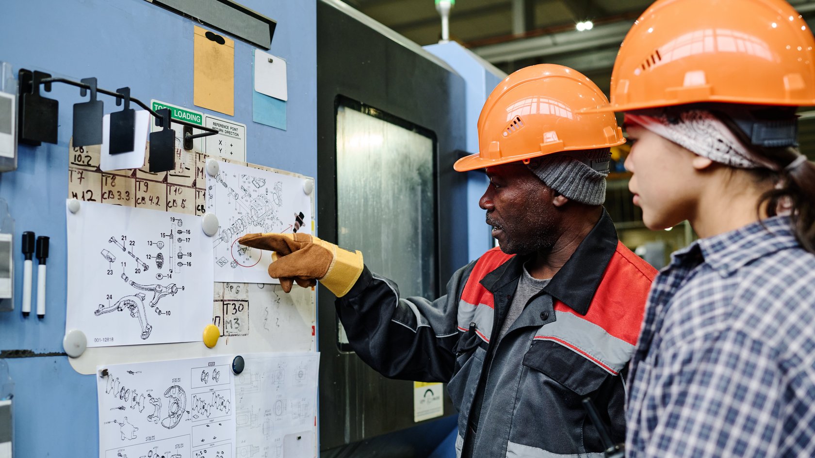 Zwei ausländische Fachkräfte untersuchen detaillierte, an eine Pinnwand gepinnte Baupläne und diskutieren wichtige Aspekte der technischen Zeichnungen in einem industriellen Umfeld