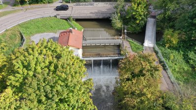 Blick von unterstrom auf das Hühnerlochwehr in der Mitte des Bildes. Hier stürzt das Wasser der Pfinz in den Pfinzentlastungskanal. Links des Wehrs ist das Betriebshaus, oberhalb des Wehrs ein Bediensteg und rechts ein kleiner Steg und eine weiße Brücke. Die weiße Brücke ist der Fuß- und Radweg. Der kleine Steg ist der Betriebssteg über die Pfinzschütze. Die Pfinz kommt von oben und fließt nach rechts. Oben im Bild ist die Eisenbahnstraße. Im Vordergrund der Pfinzentlastungskanal mit Gehölzen.