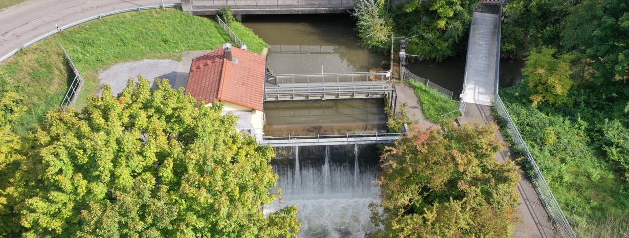 Blick von unterstrom auf das Hühnerlochwehr in der Mitte des Bildes. Hier stürzt das Wasser der Pfinz in den Pfinzentlastungskanal. Links des Wehrs ist das Betriebshaus, oberhalb des Wehrs ein Bediensteg und rechts ein kleiner Steg und eine weiße Brücke. Die weiße Brücke ist der Fuß- und Radweg. Der kleine Steg ist der Betriebssteg über die Pfinzschütze. Die Pfinz kommt von oben und fließt nach rechts. Oben im Bild ist die Eisenbahnstraße. Im Vordergrund der Pfinzentlastungskanal mit Gehölzen.