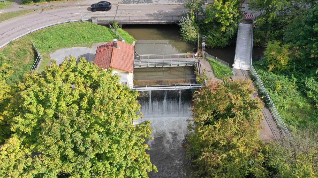 Blick von unterstrom auf das Hühnerlochwehr in der Mitte des Bildes. Hier stürzt das Wasser der Pfinz in den Pfinzentlastungskanal. Links des Wehrs ist das Betriebshaus, oberhalb des Wehrs ein Bediensteg und rechts ein kleiner Steg und eine weiße Brücke. Die weiße Brücke ist der Fuß- und Radweg. Der kleine Steg ist der Betriebssteg über die Pfinzschütze. Die Pfinz kommt von oben und fließt nach rechts. Oben im Bild ist die Eisenbahnstraße. Im Vordergrund der Pfinzentlastungskanal mit Gehölzen.