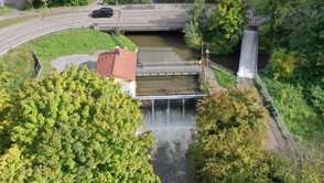 Blick von unterstrom auf das Hühnerlochwehr in der Mitte des Bildes. Hier stürzt das Wasser der Pfinz in den Pfinzentlastungskanal. Links des Wehrs ist das Betriebshaus, oberhalb des Wehrs ein Bediensteg und rechts ein kleiner Steg und eine weiße Brücke. Die weiße Brücke ist der Fuß- und Radweg. Der kleine Steg ist der Betriebssteg über die Pfinzschütze. Die Pfinz kommt von oben und fließt nach rechts. Oben im Bild ist die Eisenbahnstraße. Im Vordergrund der Pfinzentlastungskanal mit Gehölzen.