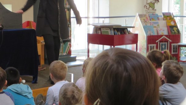 Blick von hinten auf eine Kindergruppe. Die Kinder schauen auf ein Kamshibai, ein japanisches Koffertheater, in welchem eine Bildszene aus dem Kinderbuch „Pippilothek??? Eine Bibliothek wirkt Wunder“ von Lorenz Pauli und Kathrin Schärer zu sehen ist. 