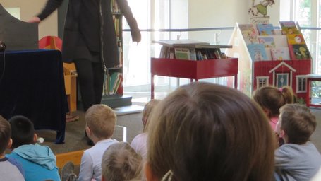 Blick von hinten auf eine Kindergruppe. Die Kinder schauen auf ein Kamshibai, ein japanisches Koffertheater, in welchem eine Bildszene aus dem Kinderbuch „Pippilothek??? Eine Bibliothek wirkt Wunder“ von Lorenz Pauli und Kathrin Schärer zu sehen ist. 