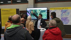 Einige Menschen betrachten einen Monitor. Jemand zeigt etwas darauf. Man sieht farbige Linien und Flächen in einem Landschaftsbild. Die daneben hängenden Pläne zeigen hydraulische Berechnungen der Wassertiefe. 