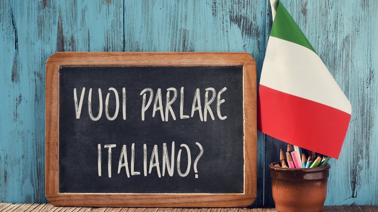 Kreidetafel mit Beschriftung "vuoi parlare italiano?" vor türkisfarbener Wand. Davor ein Gefäß mit Stiften und spanischer Fahne. Rechts neben dem Gefäß liegen zwei übereinander gestapelte Bücher. 