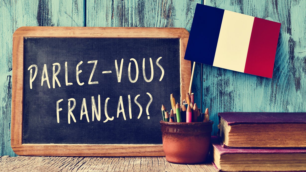 Kreidetafel mit Beschriftung "parlez-vous francais?" vor türkisfarbener Wand. Davor ein Gefäß mit Stiften und spanischer Fahne. Rechts neben dem Gefäß liegen zwei übereinander gestapelte Bücher. 