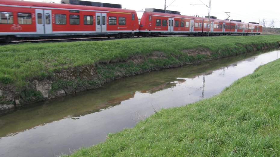 Bereich Wertstoffhof AVR - Blick in Fließrichtung Auf dem Bild sieht man im Vordergrund den monotonen Gewässerlauf des Leimbachs und im Hintergrund die unmittelbar angrenzende parallel verlaufende Bahntrasse auf der gerade ein Zug fährt.