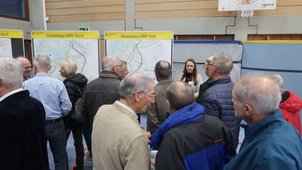 Einige Menschen betrachten die Pläne, die ausgehängt sind und sprechen miteinander. Die Überschrift des betrachteten Plans lautet „Dammbau“. 