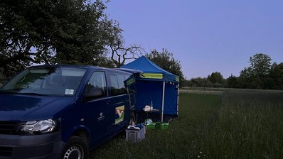 In der Dämmerung parkt auf einer Wiese ein dunkelblauer Kleinbus mit Pavillon. An der Fahrzeugseite ist die Aufschrift Ökomobil und eine Fledermaus zu erkennen.