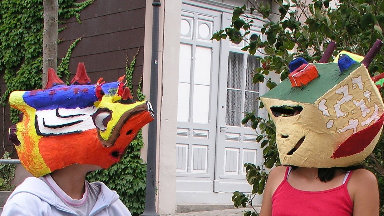 Schülerin und Schüler im Kunstunterricht, die sich gegenübersitzend ansehen und dabei selbst gestaltete bunte Masken tragen