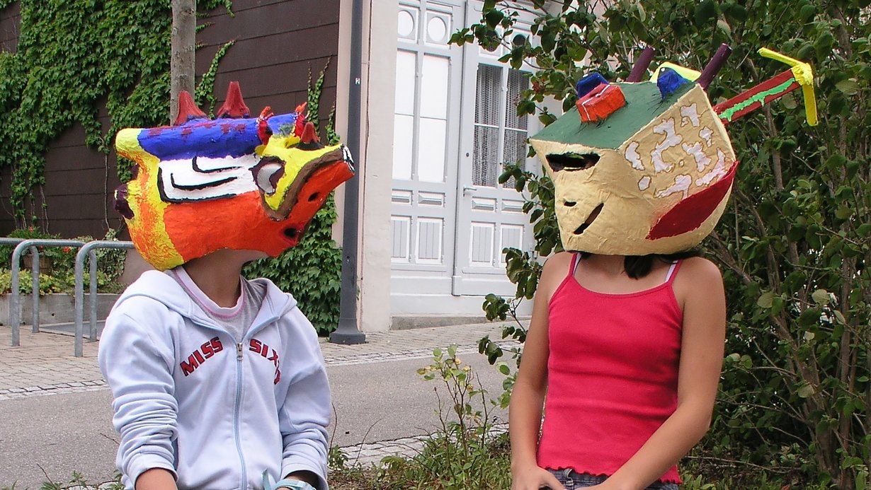 Schülerin und Schüler im Kunstunterricht, die sich gegenübersitzend ansehen und dabei selbst gestaltete bunte Masken tragen