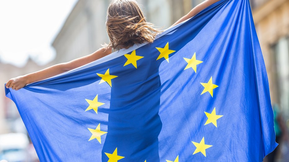 Junges Mädchen winkt mit der Flagge der Europäischen Union im Rücken in der Stadt.