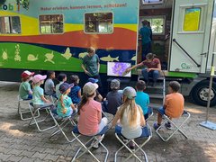 Eine Gruppe von Kindern sitzt im Halbkreis neben dem Ökomobil und schaut auf einen Bildschirm, neben dem sich die Ökomobilistinnen befinden. Gezeigt wird eine lila Blüte in Nahaufnahme.