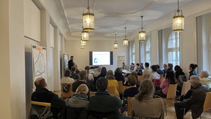 Auf dem Foto sieht man ca. 30 Personen in einem Besprechungsraum. Der Projektleiter erläutert die Hydraulik des Projekts anhand einer Powerpoint-Präsentation. Pläne hängen an Stellwänden aus. 