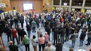 Eine Turnhalle mit ca. 200 Personen. Es wird ein Vortrag gehalten. An den Wänden sieht man Stellwände, an denen Pläne aushängen. 