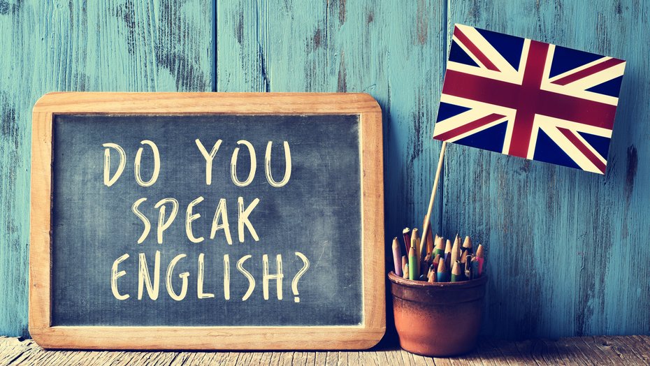 Kreidetafel mit Beschriftung "do you speak english?" Kreidetafel mit Beschriftung "do you speak english?" vor türkisfarbener Wand. Davor ein Gefäß mit Stiften und spanischer Fahne. Rechts neben dem Gefäß liegen zwei übereinander gestapelte Bücher.