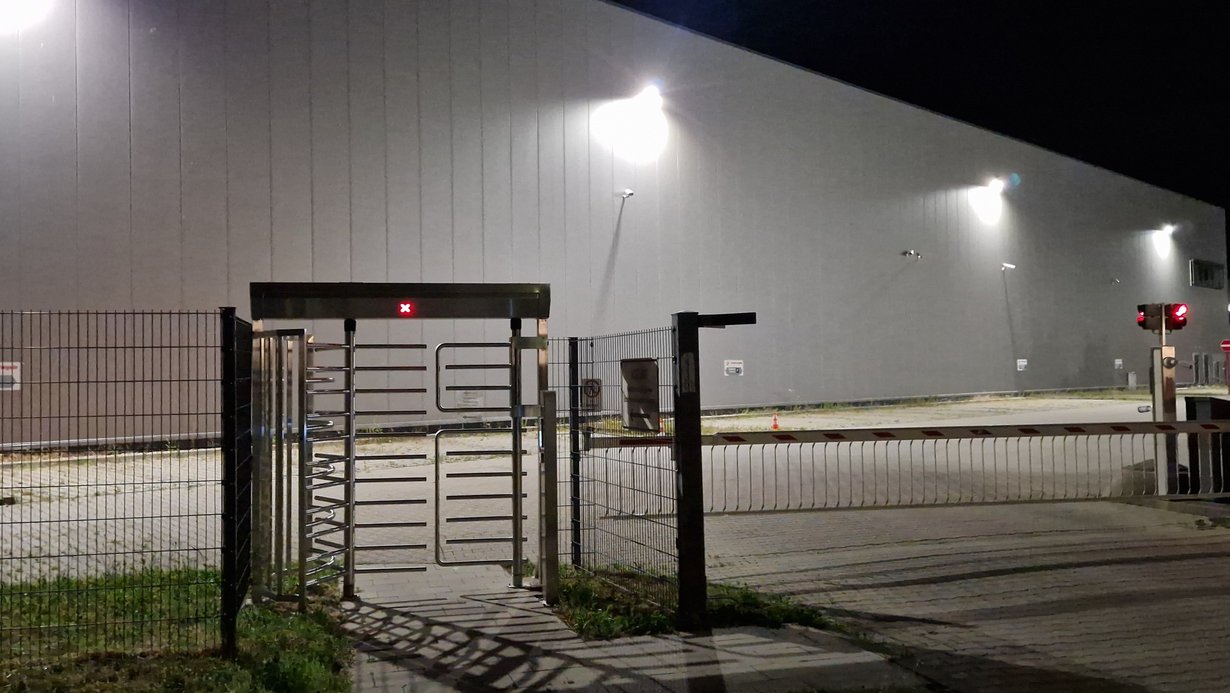 Hell erleuchtete Fassade einer Lagerhalle mit Schranke und Drehkreuz im Vordergrund