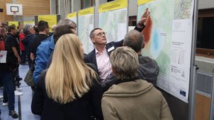 Einige Menschen betrachten die Pläne, die ausgehängt sind. Jemand zeigt etwas im Plan. Die Überschrift der Pläne im Hintergrund lauten „Schutzmaßnahmen“. 