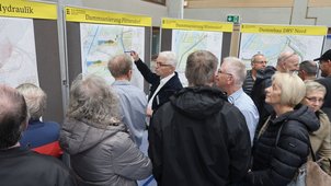 Einige Menschen betrachten die Pläne, die ausgehängt sind. Jemand zeigt etwas im Plan. Die Überschrift des betrachteten Plans lautet „Dammsanierung Plittersdorf“. 