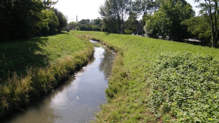 Bereich Brücke in den Weinäckern - Blick gegen Fließrichtung Auf dem Bild sieht man den monotonen und von seitlichen Dammstrukturen eingeengten Leimbach. Die Ufer und Dammbereiche sind mit Gras bewachsen. Auf beiden Seiten sieht man im Anschluss Bäume und Sträucher. Auf der rechten Seite erkennt man im Hintergrund eine Straße mit Straßenschild. In der Ferne sieht man eine Brücke, einen Turm sowie einen Kran.