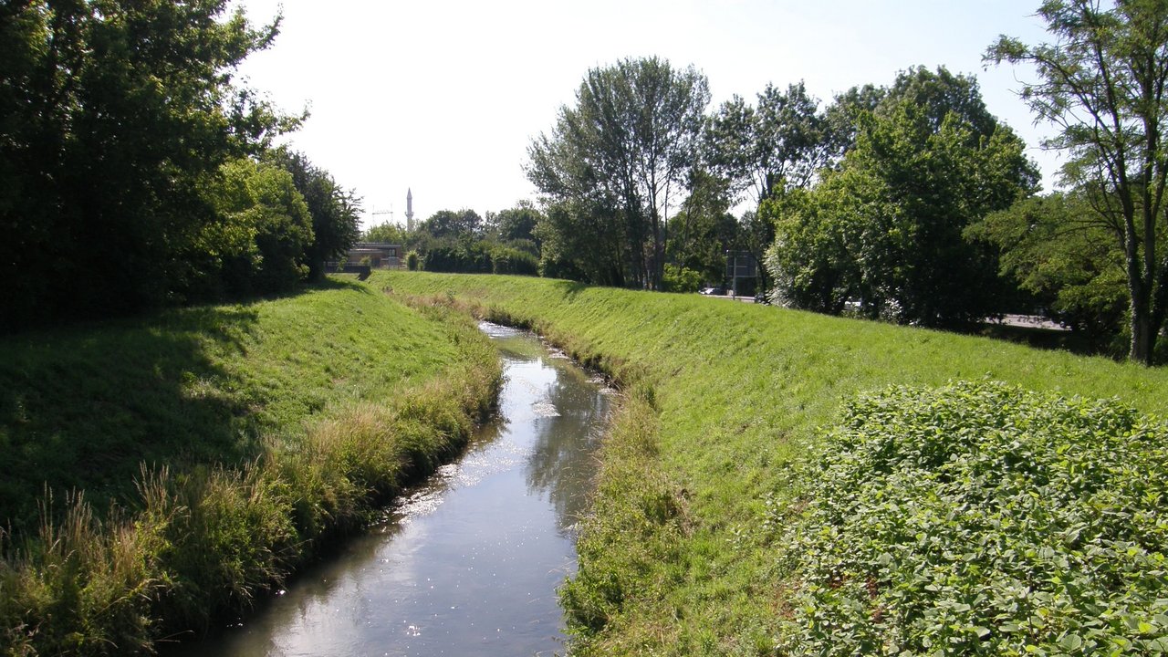 Auf dem Bild sieht man den monotonen und von seitlichen Dammstrukturen eingeengten Leimbach. Die Ufer und Dammbereiche sind mit Gras bewachsen. Auf beiden Seiten sieht man im Anschluss Bäume und Sträucher. Auf der rechten Seite erkennt man im Hintergrund eine Straße mit Straßenschild. In der Ferne sieht man eine Brücke, einen Turm sowie einen Kran.
