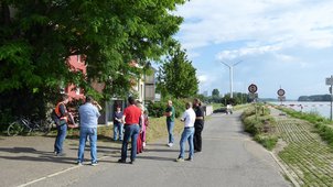 Auf dem Foto sieht man den Projektleiter im Gespräch mit 10 Bürgerinnen und Bürgern am Damm auf Höhe der Wohnhäuser im Juli 2021. Er zeigt ihnen Pläne und erläutert diese. 