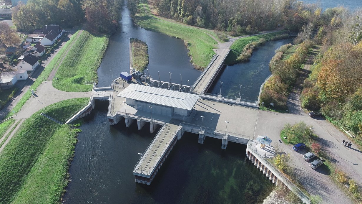 Es wird das Acherschöpfwerk als Luftaufnahme mittels Drohne gezeigt. Durch das auf rechter Bildseite daneben liegende dreizügige Durchlassbauwerk F besteht die Fließverbindung vom Teilpolder 2 in den Rheinseitengraben. Die Acher fließt außerhalb der Hochwasserzeiten am linken Bildrand durch das Siel in den Rheinseitengraben.