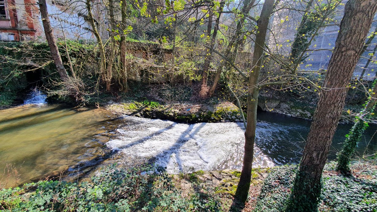 Die Weschnitz fließt von links nach rechts. Der kurze steile Abschnitt der Weschnitz ist die sogenannten Gleite. Auf weniger als 2 Meter fällt die Sohle der Weschnitz einen Meter nach unten. Ober- und Unterstrom der Gleite befinden sich die Auslässe der angrenzenden Wasserkraftanlage. Die Ufer der Weschnitz sind mit Wasserbausteinen gesichert. Im Vorland stehen Bäume.