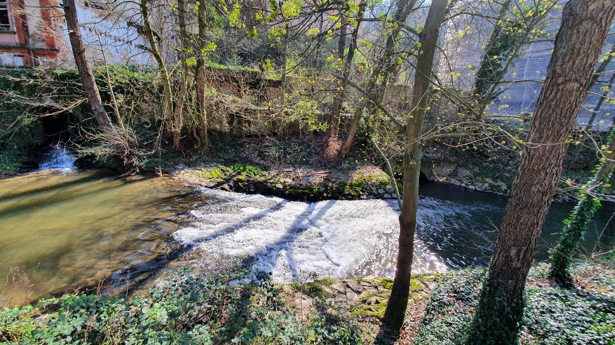 Die Weschnitz fließt von links nach rechts. Der kurze steile Abschnitt der Weschnitz ist die sogenannten Gleite. Auf weniger als 2 Meter fällt die Sohle der Weschnitz einen Meter nach unten. Ober- und Unterstrom der Gleite befinden sich die Auslässe der angrenzenden Wasserkraftanlage. Die Ufer der Weschnitz sind mit Wasserbausteinen gesichert. Im Vorland stehen Bäume.