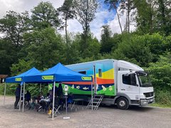Vor dem Ökomobil stehen auf einem Parkplatz zwei Pavillons mit Tischen und Stühlen. Kinder schauen sitzend durch Mikroskope.