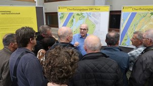 Einige Menschen betrachten die Pläne, die ausgehängt sind. Die Überschrift des Plans lautet „Überflutungsdauer“. Ein Mann spricht zu der Gruppe und erläutert den Plan. 