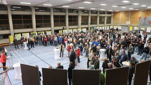 Eine Turnhalle mit ca. 200 Personen. Es wird ein Vortrag gehalten. An den Wänden sieht man viele Stellwände, an denen Pläne aushängen. 