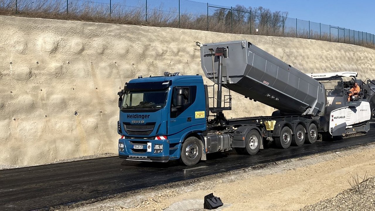 Zu sehen ist die B 292 Ortsumgehung Adelsheim im Bau. Genauer: Zwischen der neuen Spritzbeton-Hangsicherung links und einer Einschnittsböschung rechts baut eine Asphaltkolonne die neue Fahrbahn ein. Im Vordergrund entlädt ein LKW Asphalt in den Beschicker. Dahinter folgen der Asphalt-Fertiger und zwei Walzen.