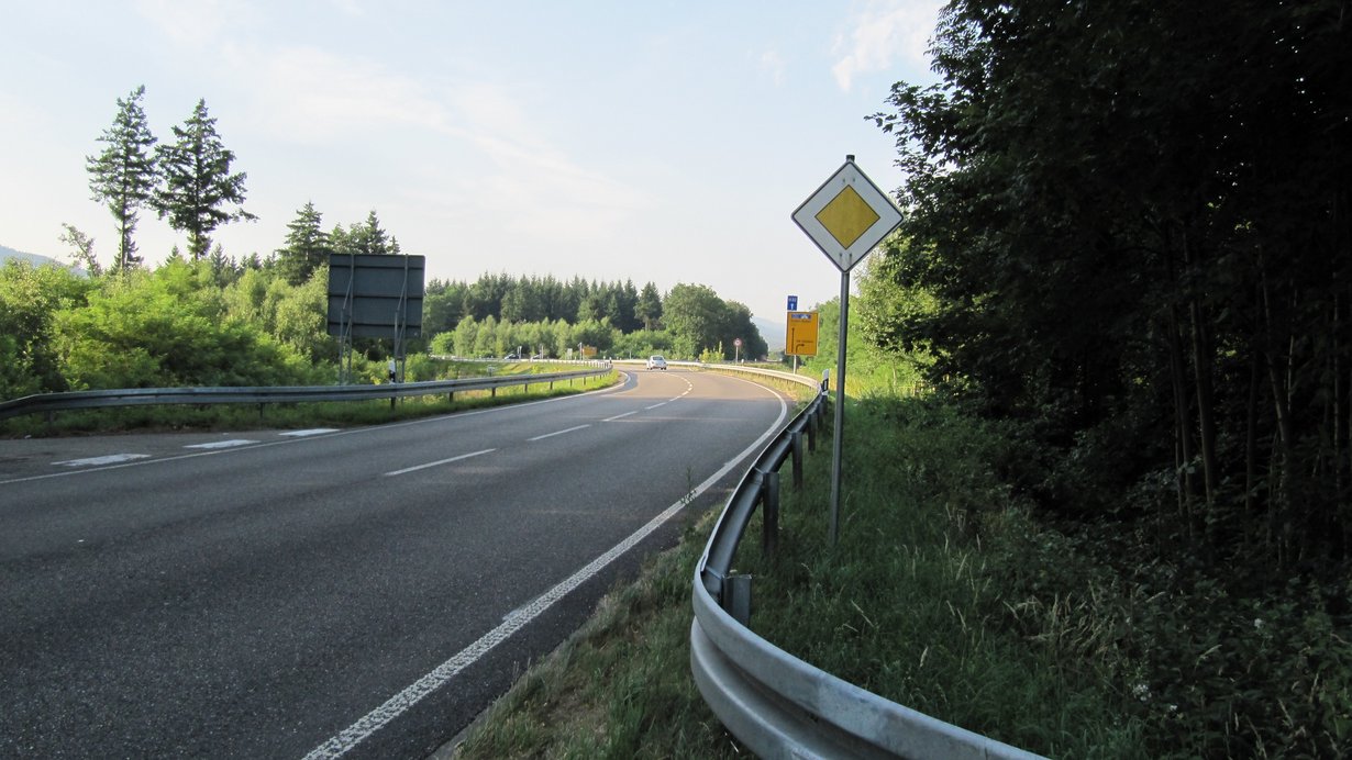 Jetzige Führung der B3 (Blickrichtung Süden); im Hintergrund fährt ein kleines weißes Auto; im Bereich dieses Fahrzeugs beginnt künftig der Straßenneubau; im Vordergrund ein Vorfahrtsschild, dahinter ein gelbes Verkehrsschild, das die Fahrtrichtung Autobahn sowie den Abzweig Richtung Sandweier zeigt. Auto; im Vordergrund ein Vorfahrtsschild, dahinter ein gelbes Verkehrsschild, dahinter ein gelbes Verkehrsschild, das die Fahrtrichtung Autobahn zeigt.