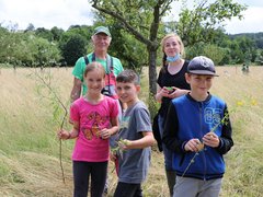 Eine Ökomobilistin steht gemeinsam mit dem Veranstalter und drei Kindern auf einer Wiese und alle lächeln in die Kamera. Die Kinder halten ihre gesammelten Pflanzen in den Händen. 