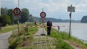 Das Foto ist entlang des Rheinhochwasserdamms aufgenommen. Der Blick geht von der Rheinbrücke in Richtung Hofgut Maxau. Man sieht, dass aufgrund des anstehenden Hochwassers der Damm mit Warnschildern und rot-weißem Band abgesperrt ist. Das Betreten ist verboten. Es besteht Lebensgefahr. Trotzdem sind mehrere Menschen auf dem Damm unterwegs und nutzen die dortigen Bänke für die Aussicht auf den Rhein bei Hochwasser. Ein Mann lehnt vorn an der Absperrung und hält sich daran, sie nicht zu überqueren. 