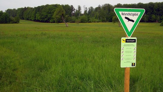 Reich strukturierte Landschaft mit Wiesen, Hecken, Streuobstbäumen und Wald. Im Vordergrund steht das dreieckige Schild mit dem Naturschutzgebiete im Gelände beschildert werden. 